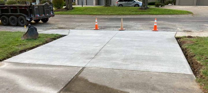 A newly constructed concrete driveway in a residential neighborhood. The poured concrete is smooth and light gray in color, with expansion joints. There are houses with driveways and yards in the background.