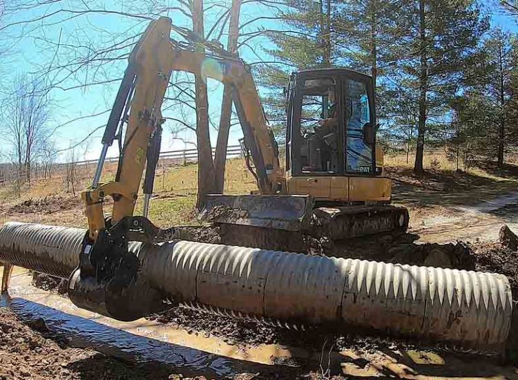 A bulldozer pushing a large metal pipe through muddy ground for drainage solutions.
